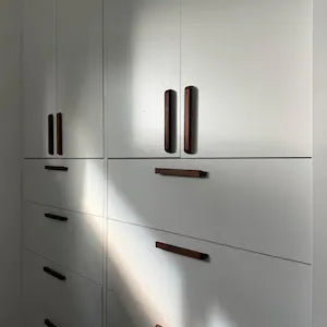 White modern wardrobe or closet doors and drawers with dark brown vertical and horizontal handles, shown in a real interior setting with sunlight casting shadows.
