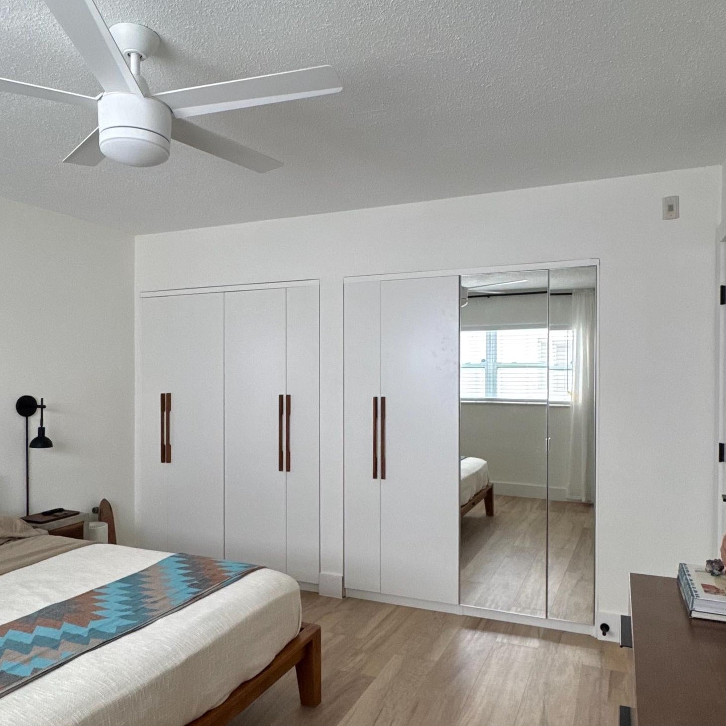 A modern bedroom interior with a wooden bed frame, white bedding, white walls, light wood flooring, a ceiling fan, and a built-in wardrobe. An open doorway reveals another room in the background.